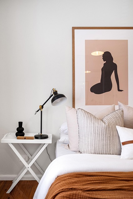 morceau d'un lit avec oreillers clair , cadre femme noir sur fond rose sur le mur, et chevet blanc avec lampr archi noire
